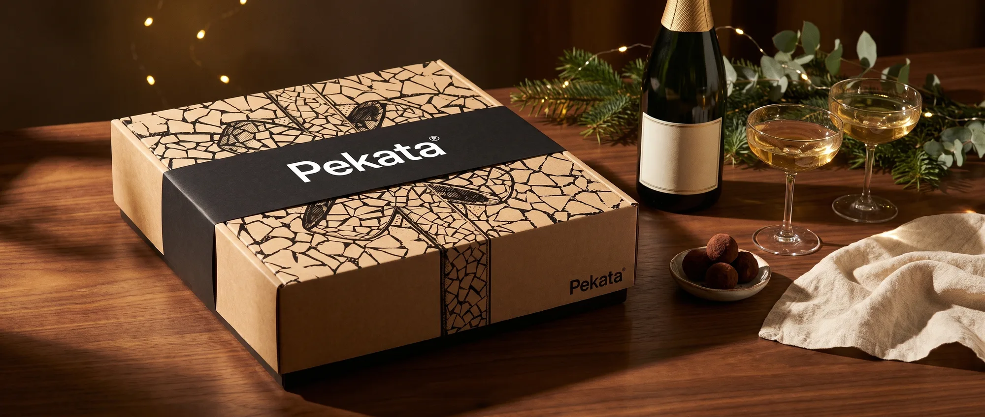 Boîte de Noël Pekata fermée avec bande noire à côté d'une bouteille de cava et deux verres sur une table en noyer